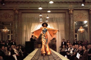a woman in a bright orange dress walks down a runway with her arms open wide as an audience watches her